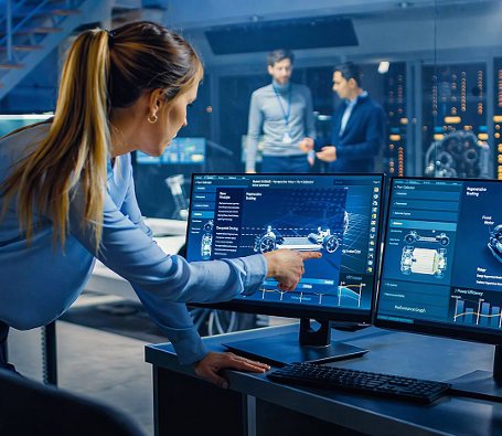 An engineer pointing at a design on two monitors in a control room, colleagues behind her
